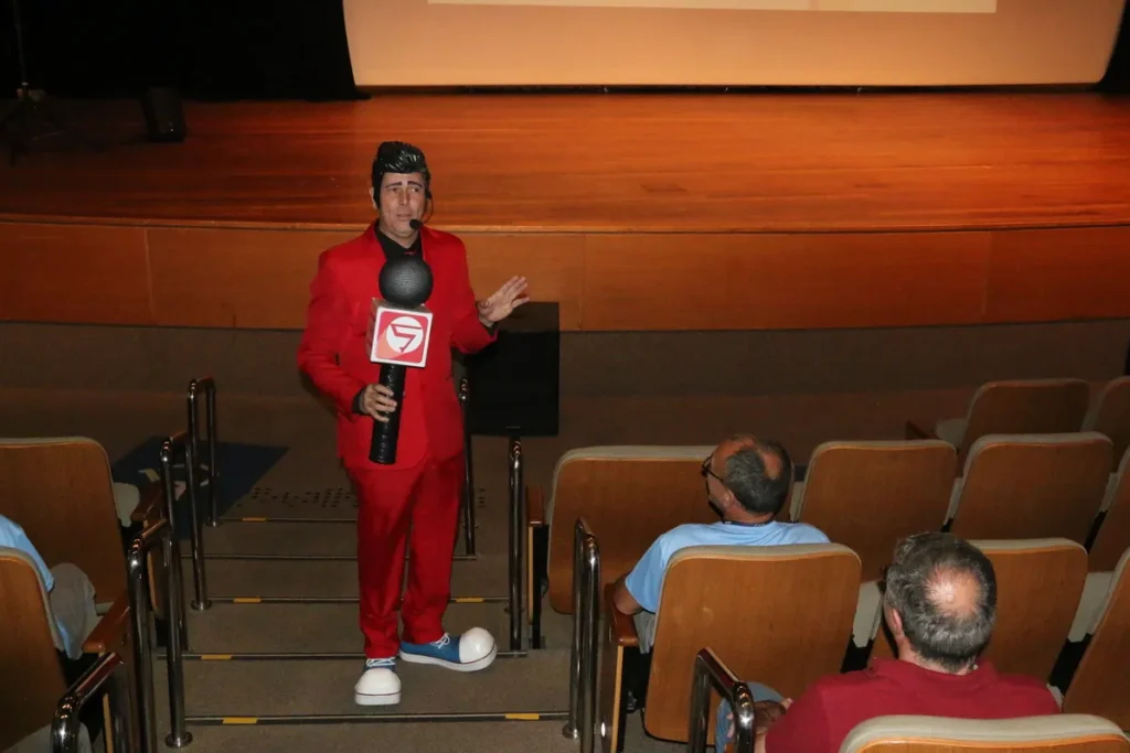 Homem vestido com terno vermelho e em estilo de personagem (Cipeiro), segurando um grande microfone preto e vermelho com o logo "S", em um auditório, gesticulando para um pequeno público, ideal para ilustrar a Análise Preliminar de Risco (APR).
