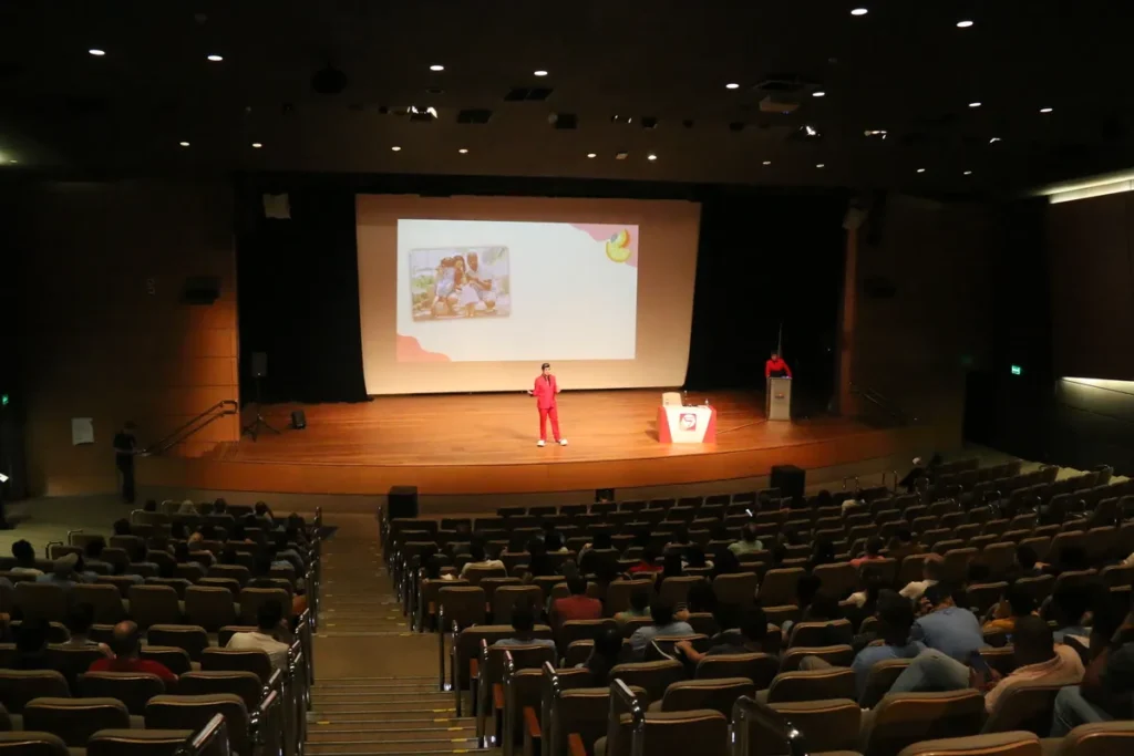 Pessoa palestrando sobre amamentação no palco de um auditório, com tela de projeção e público assistindo.