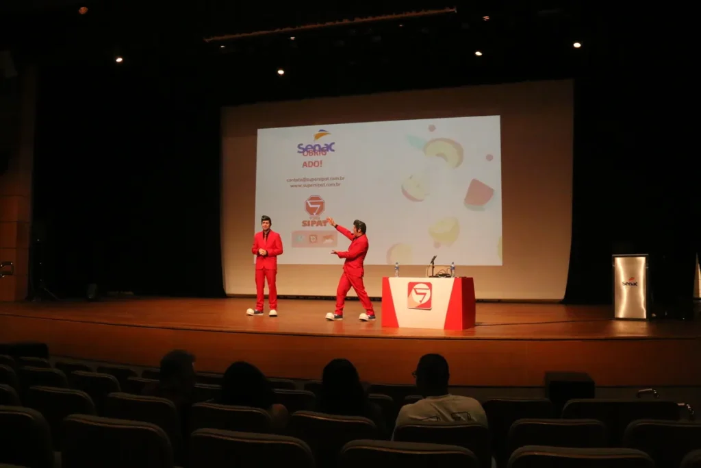 Dois palestrantes em roupas vermelhas no palco de um auditório durante uma palestra sobre amamentação.