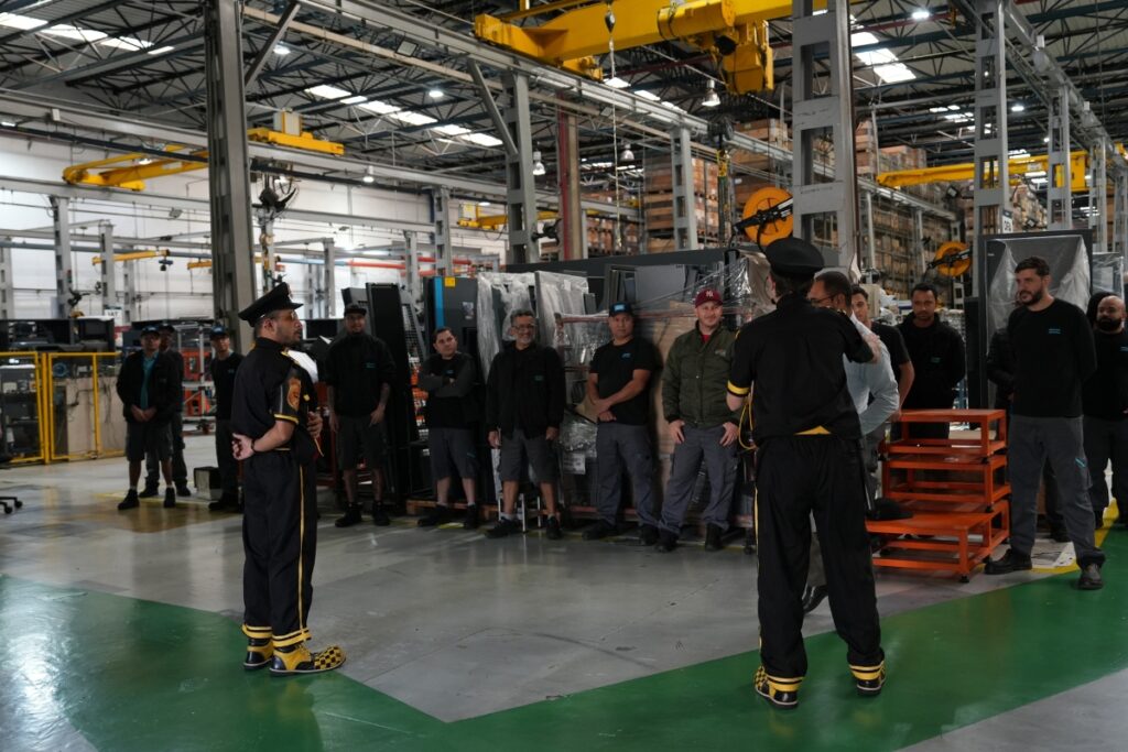 Um grupo de trabalhadores de fábrica em uniformes escuros reunido em um grande galpão industrial, ouvindo dois palestrantes com uniformes pretos e detalhes amarelos, um deles calçando sapatos de palhaço amarelos e pretos.