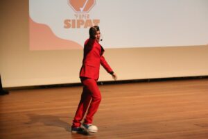 Fotografia em plano inteiro mostrando um homem de terno vermelho e sapatos coloridos se apresentando dinamicamente em um palco de madeira, diante de um telão que exibe o logotipo "Super SIPAT".