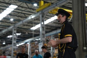 Palestrante caracterizado com uniforme preto e detalhes amarelos, usando quepe e óculos de proteção, gesticula durante apresentação em um grande galpão industrial com operários ao fundo.