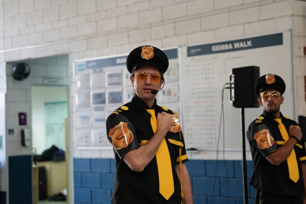 Dois palestrantes caracterizados com uniformes pretos e gravatas amarelas de inspiração policial realizam uma apresentação teatral com a mão no peito diante de um quadro de avisos com os dizeres "GEMBA WALK".