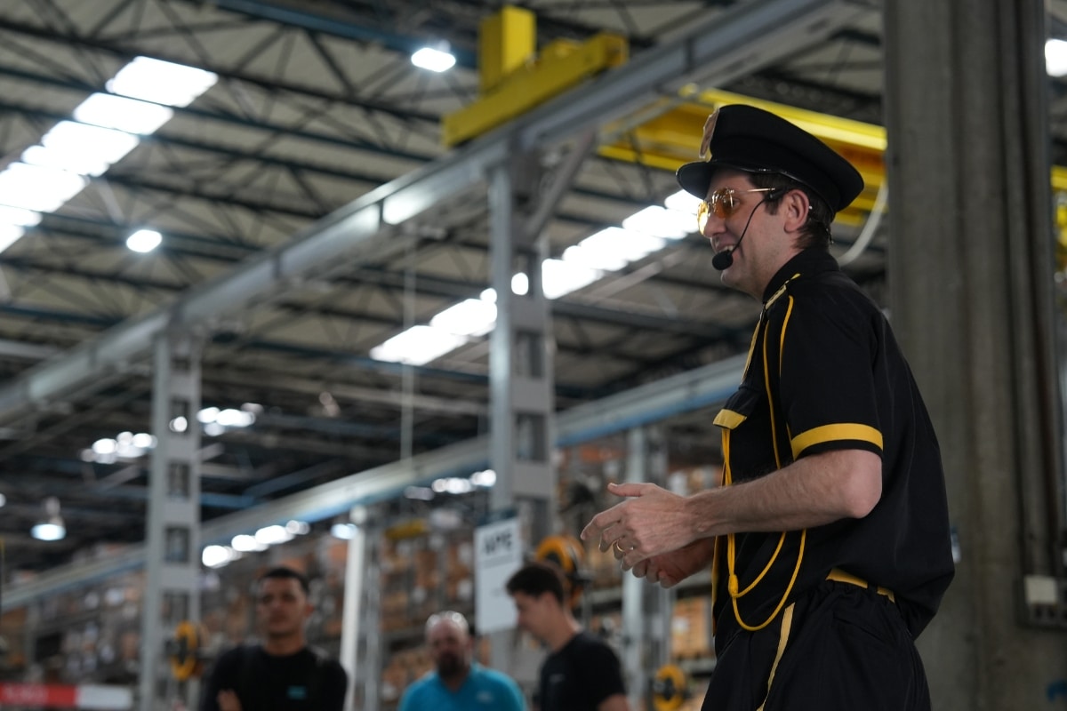 Palestrante caracterizado com uniforme preto e detalhes amarelos, usando quepe e óculos de proteção, gesticula durante apresentação em um grande galpão industrial com operários ao fundo.