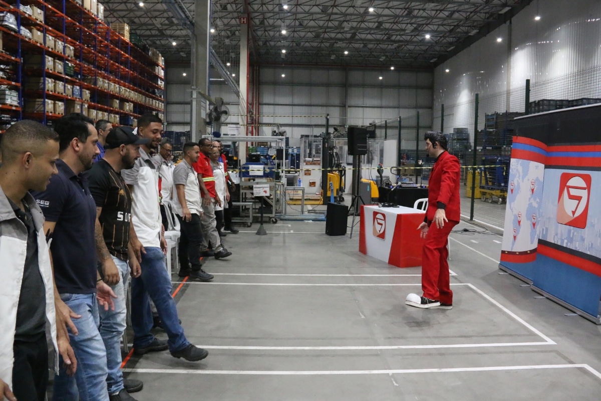 Um palestrante vestido com terno vermelho e sapatos de palhaço faz uma apresentação dinâmica para um grupo de operários em pé dentro de um grande galpão logístico ou fábrica.