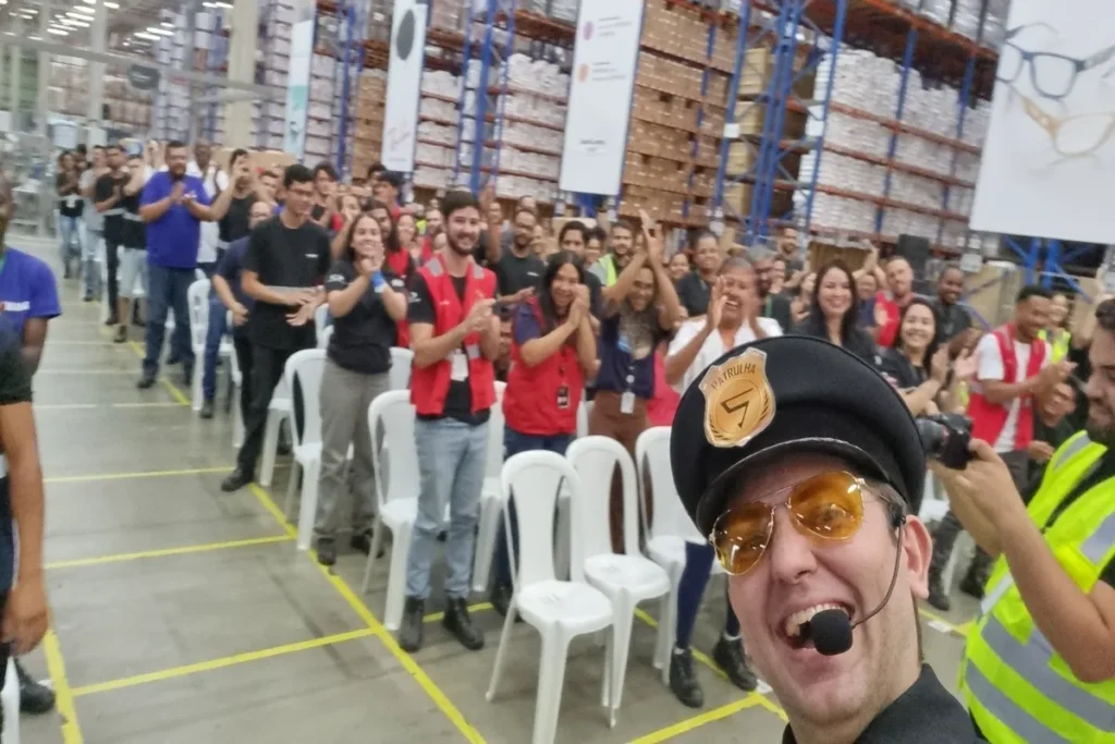 Um homem sorrindo, usando óculos de sol, um boné de segurança e uma camiseta com a inscrição "Patrulha". Ele está em um armazém com prateleiras e caixas ao fundo, e uma plateia o aplaude. A imagem representa uma palestra ou evento sobre a CAT (Comunicação de Acidente de Trabalho).
