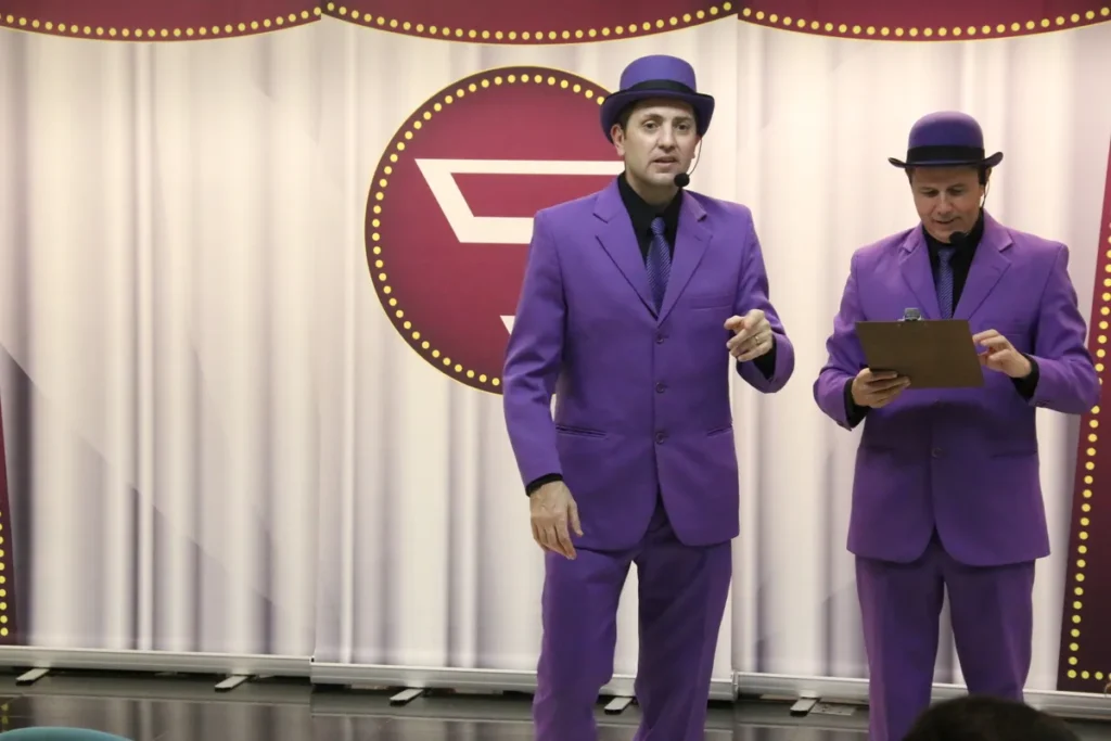 Dois homens, vestidos com ternos roxos e chapéus, em um palco. Um deles segura uma prancheta e o outro aponta, como se estivessem encenando uma peça ou uma apresentação.