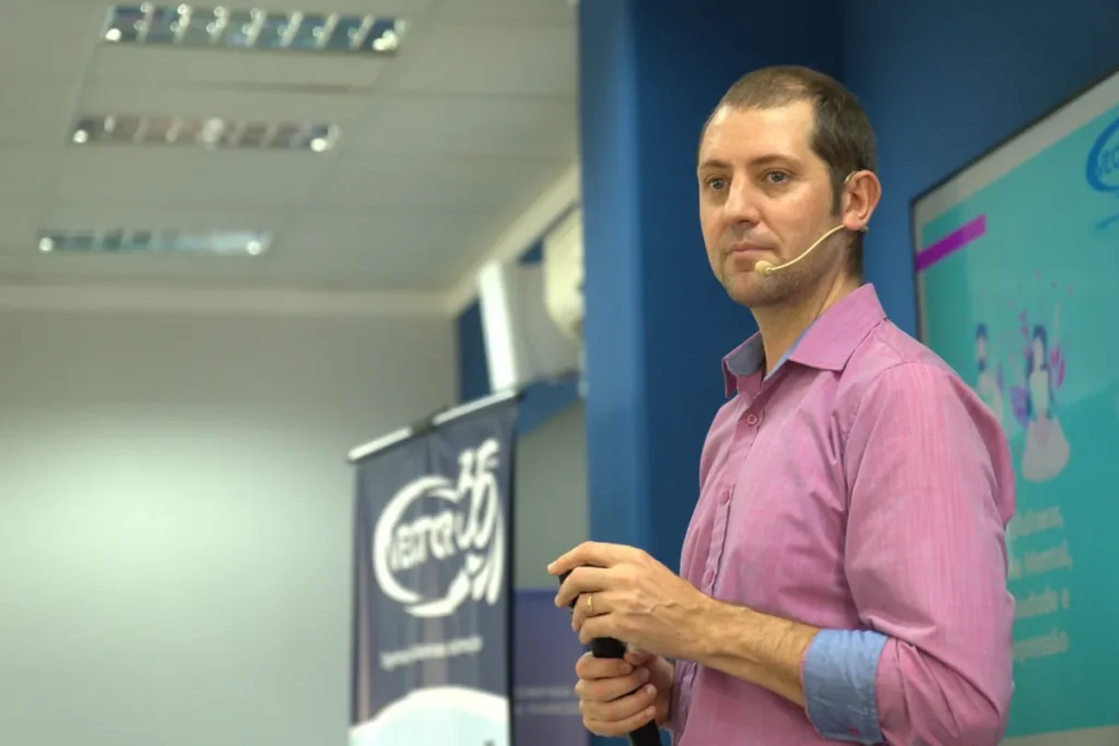 Homem palestrando em evento corporativo com microfone headset e apresentação em tela ao fundo