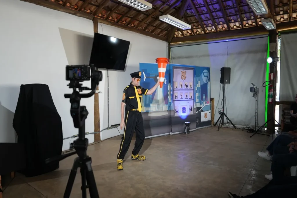 Um instrutor vestido com um uniforme temático de segurança (preto e amarelo) conduz uma dinâmica com a plateia, que está de pé com os braços estendidos. O instrutor gesticula em frente a um grande telão que exibe gráficos de prevenção de acidentes. O cenário é moderno, com uma parede de madeira e luzes neon verdes. A dinâmica ativa sugere um treinamento de primeiros socorros e a importância de uma palestra sobre DEA (Desfibrilador Externo Automático) para o ambiente de trabalho.