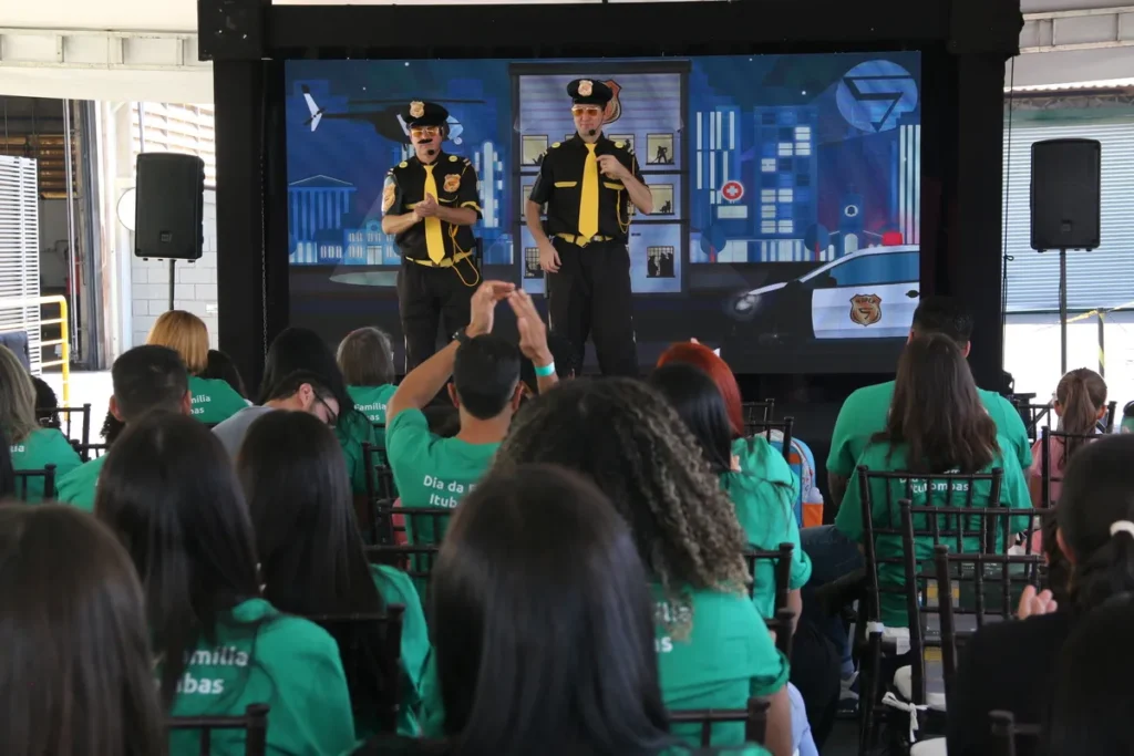 Dois homens vestidos como policiais em um palco com um telão que mostra uma paisagem urbana com um carro de polícia. Uma plateia de pessoas vestidas com camisetas verde-limão está assistindo à apresentação. A imagem representa o tema de DPOC (Doença Pulmonar Obstrutiva Crônica).