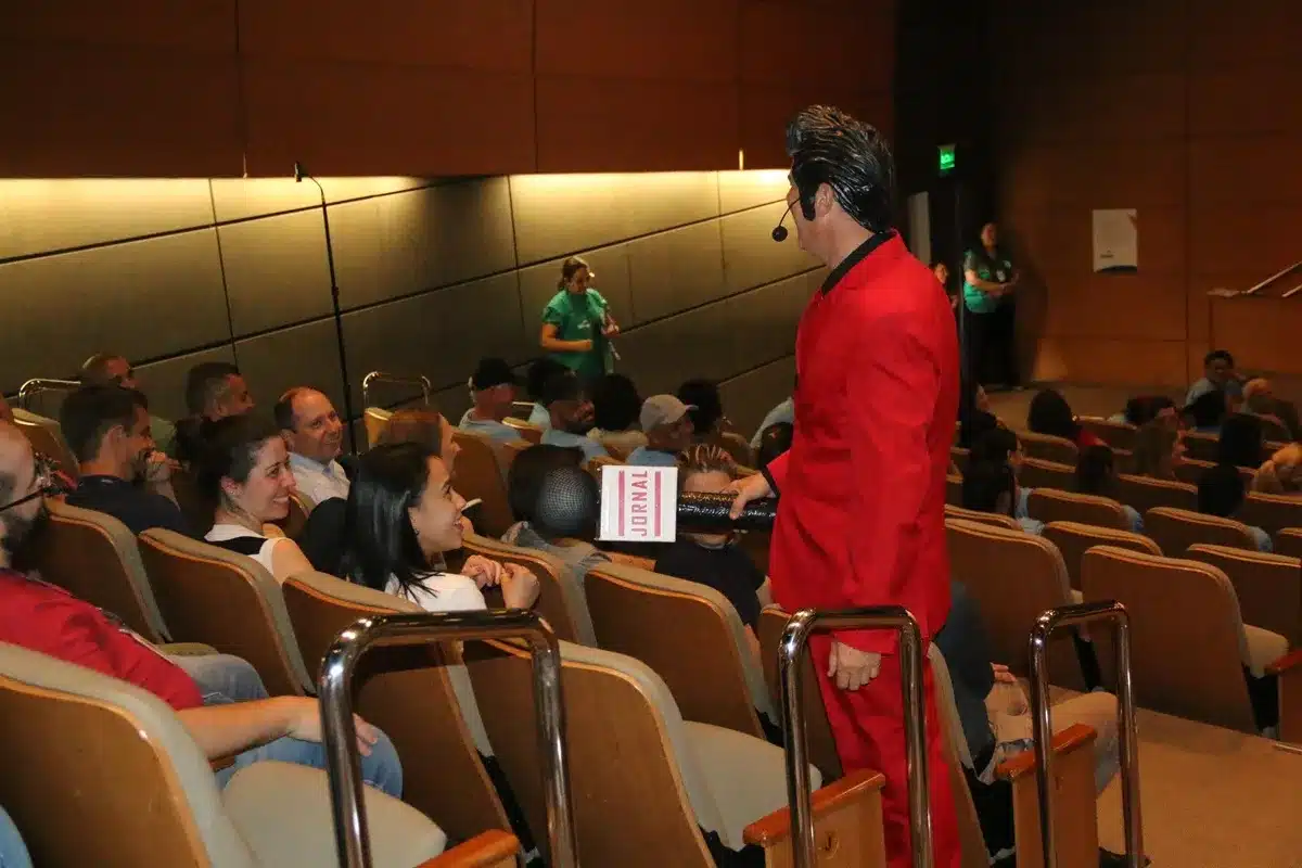 Apresentador de terno vermelho interage com o público em auditório, segurando microfone e conversando com participantes sentados.