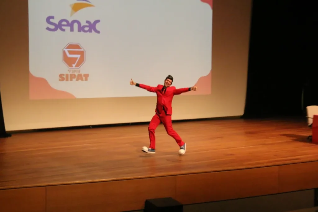 Artista em traje vermelho realiza performance teatral em palco durante evento SIPAT no Senac, com expressão animada e braços abertos diante de tela de projeção.