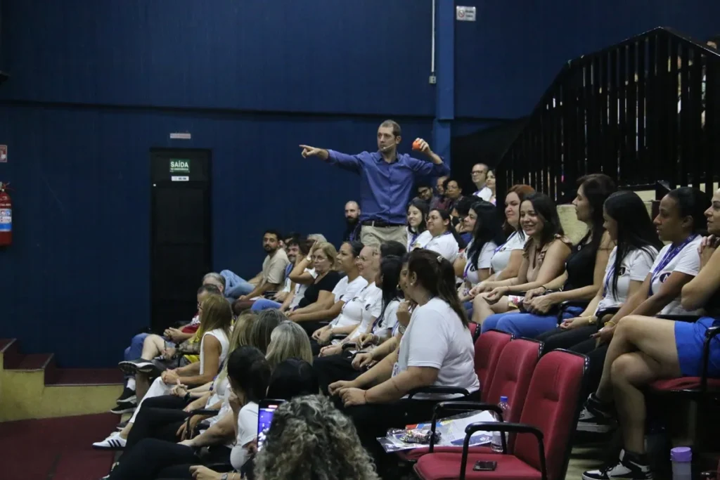 Palestrante interage com o público em auditório, apontando enquanto pessoas sentadas em cadeiras vermelhas observam e participam ativamente.