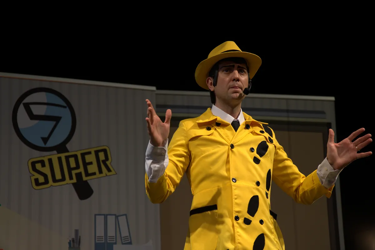 Ator vestido com terno e chapéu amarelos, usando microfone de cabeça, gesticulando durante apresentação teatral em palco com fundo preto.