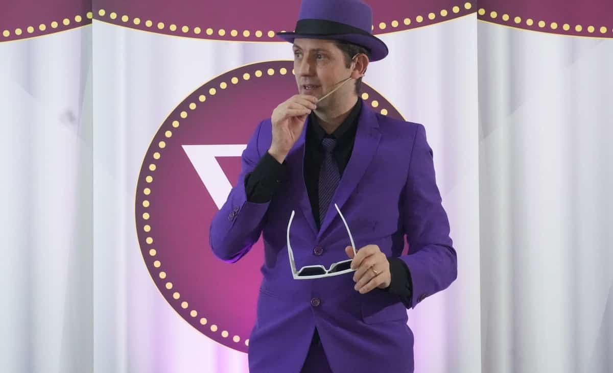 Homem usando terno e chapéu roxos, segurando óculos brancos e falando com microfone de lapela durante apresentação em evento.