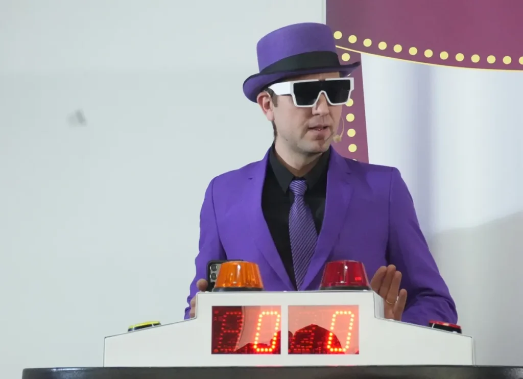 Homem vestido com terno roxo, chapéu combinando e óculos brancos grandes, em frente a um painel de pontuação digital com luzes coloridas, conduzindo uma dinâmica de perguntas em evento corporativo.
