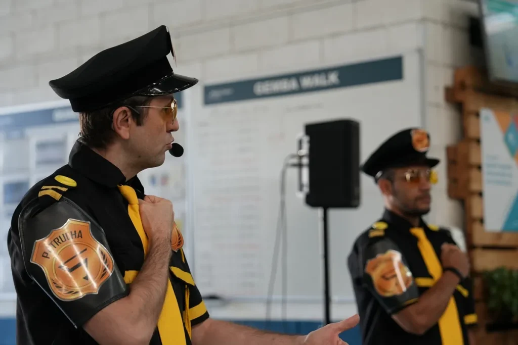 Dois palestrantes vestidos como oficiais da patrulha de segurança, com uniformes pretos e detalhes amarelos, participam de uma palestra sobre segurança no trabalho.