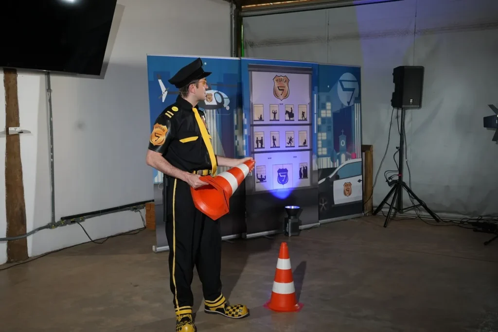 Homem vestido de policial com uniforme preto e amarelo segura dois cones de trânsito laranja em um ambiente interno decorado com painéis ilustrativos e equipamentos de som.