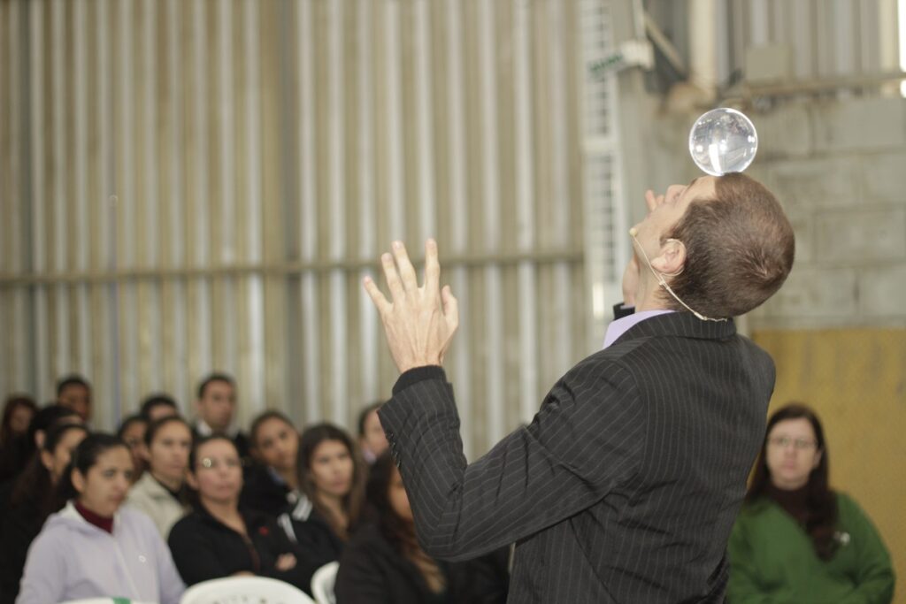 Palestrante diante de plateia equilibrando bola transparente na cabeça