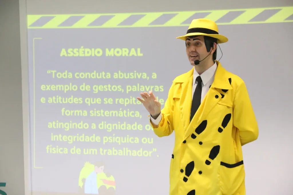 Palestrante vestido com terno amarelo e chapéu da mesma cor apresenta palestra sobre assédio moral, em frente a um slide com definição do tema.