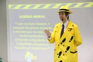 Palestrante vestido com terno amarelo e chapéu da mesma cor apresenta palestra sobre assédio moral, em frente a um slide com definição do tema.