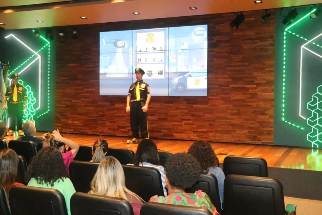 Dois atores vestidos de policiais com uniforme preto e amarelo se apresentam em um auditório diante de um público sentado, com painel digital exibindo imagens de trânsito ao fundo.