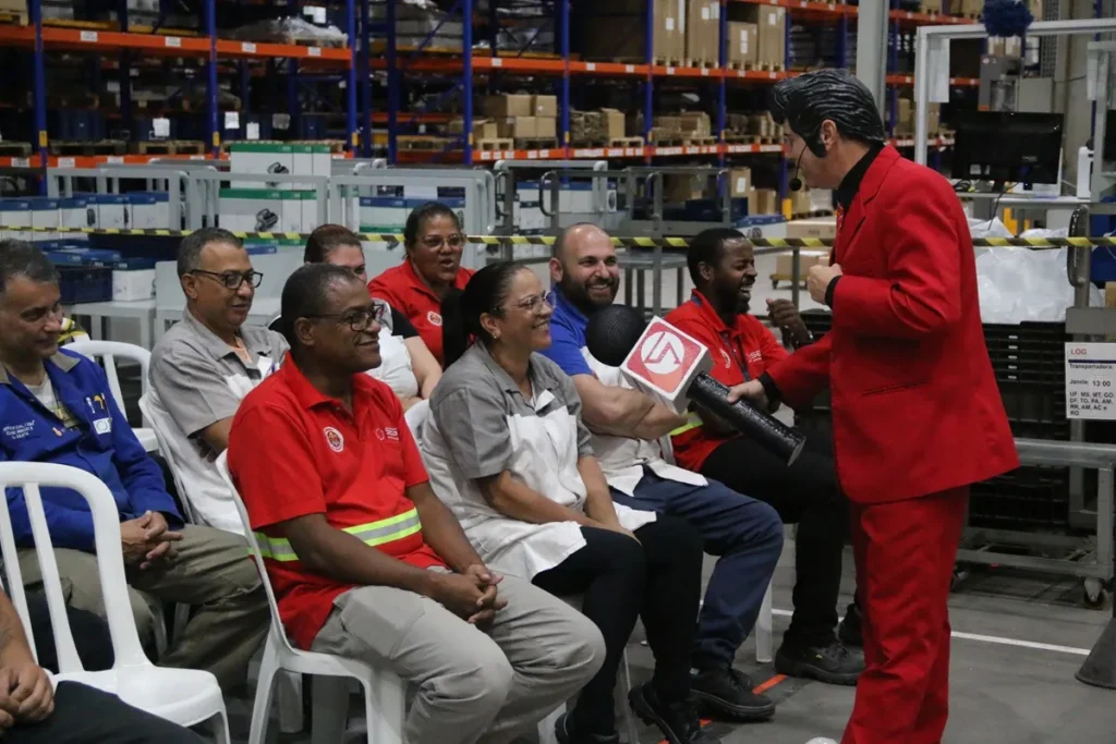 Palestrante de terno vermelho entrega um microfone gigante a colaborador sentado em cadeira plástica dentro de um galpão industrial com prateleiras de estoque ao fundo