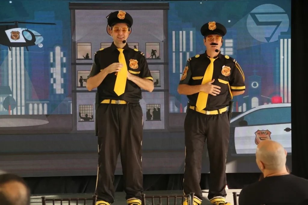 Dois apresentadores fantasiados de policiais com uniformes pretos e gravatas amarelas ministram palestra de segurança, diante de telão com cenário urbano e viatura da Super SIPAT.