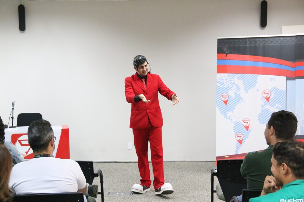 Homem de terno vermelho e microfone de cabeça interagindo com o público durante uma palestra ou apresentação corporativa.