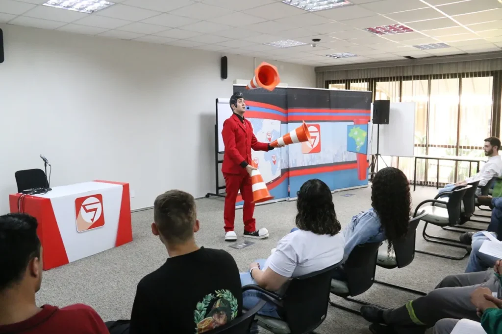 Apresentador de terno vermelho faz malabarismo com cones de trânsito laranja diante de plateia durante evento Super SIPAT.