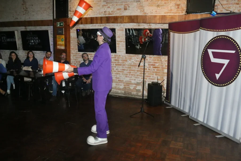 Artista vestido de roxo faz malabarismo com cones de trânsito laranja diante de uma plateia em um ambiente interno com parede de tijolos e cortina decorativa.