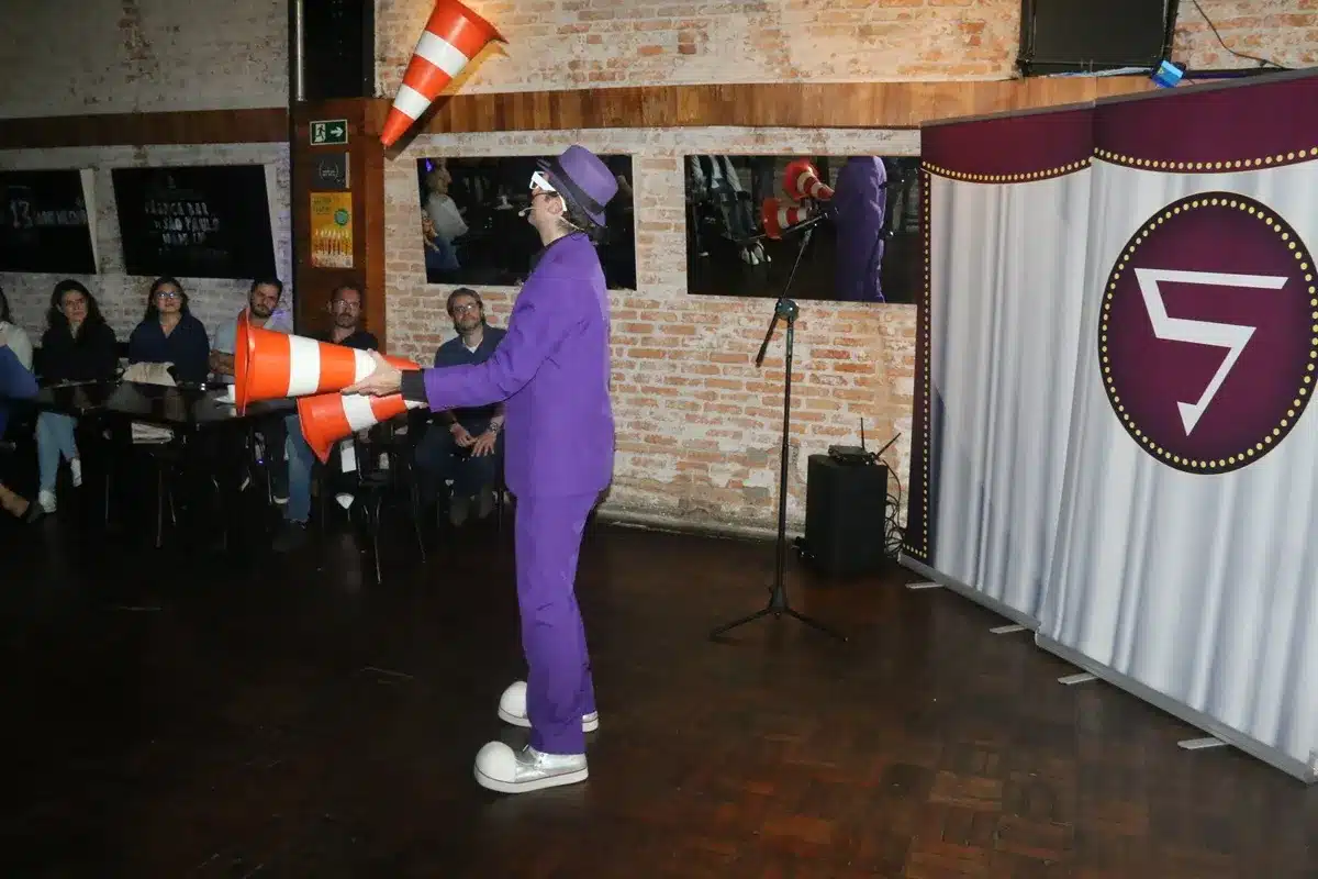 Artista vestido de roxo faz malabarismo com cones de trânsito laranja diante de uma plateia em um ambiente interno com parede de tijolos e cortina decorativa.