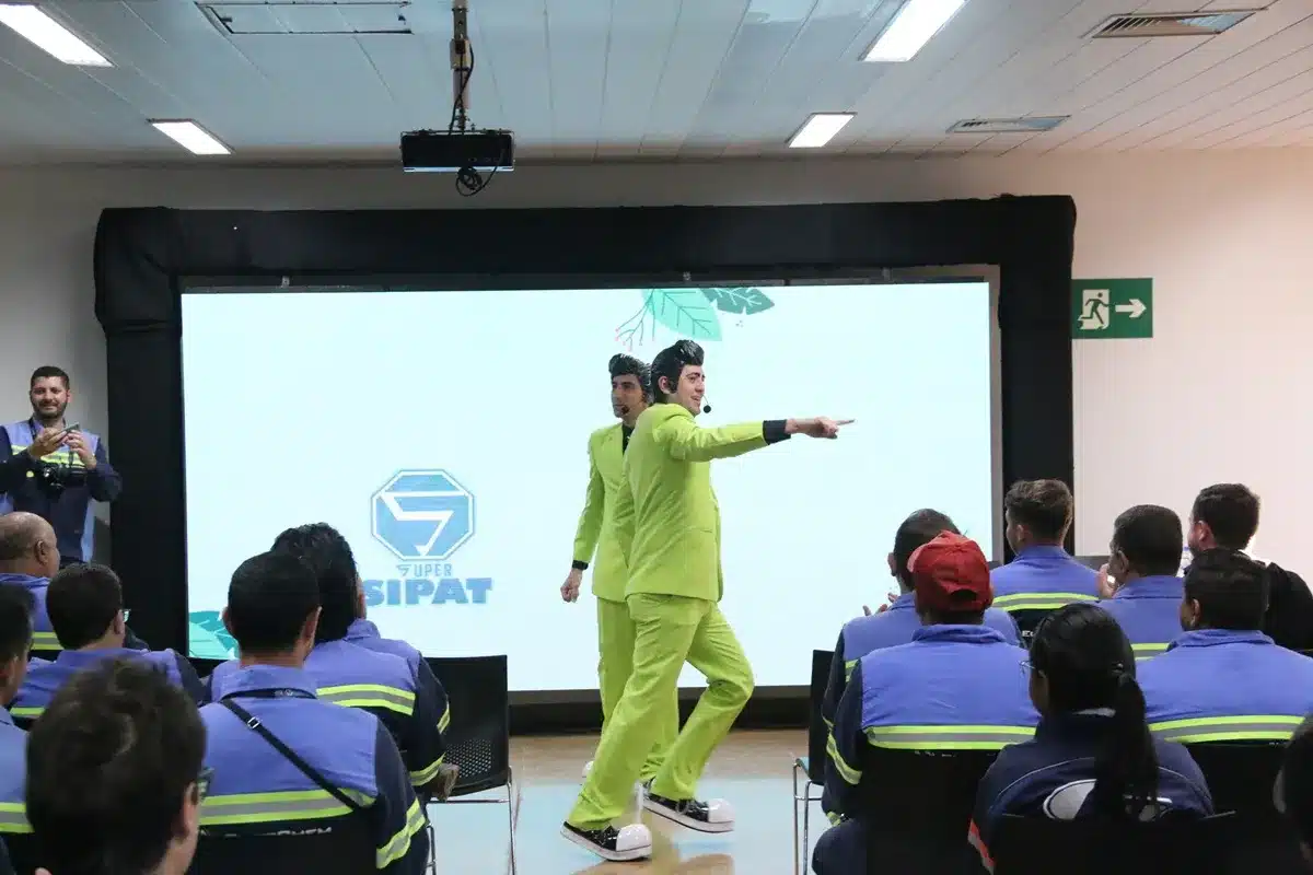 Dois artistas vestidos com ternos verdes e sapatos grandes brancos se apresentam diante de colaboradores uniformizados durante o evento SIPAT, promovendo conscientização sobre segurança de forma divertida e interativa.