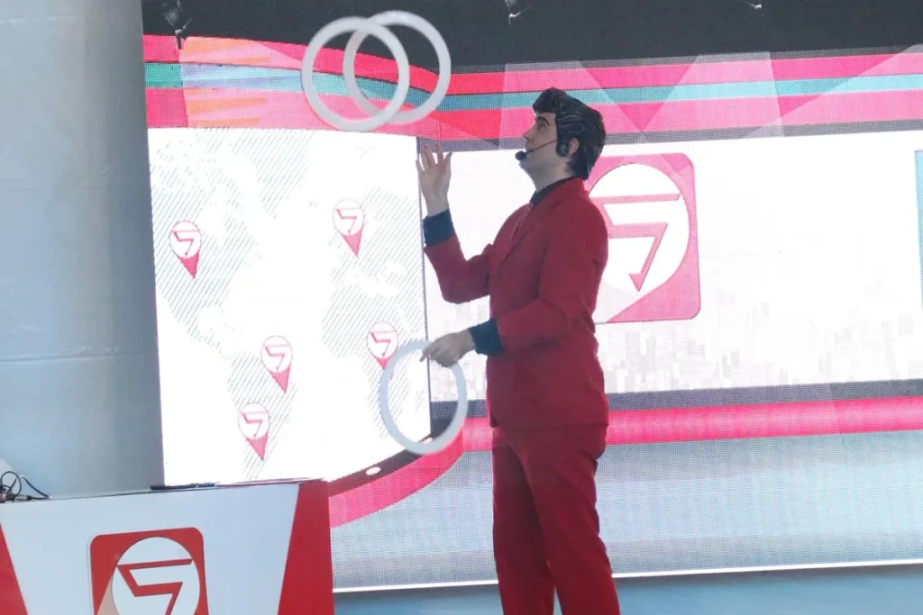 Homem vestido com terno vermelho realizando malabarismo com argolas brancas durante apresentação em evento com telão ao fundo.