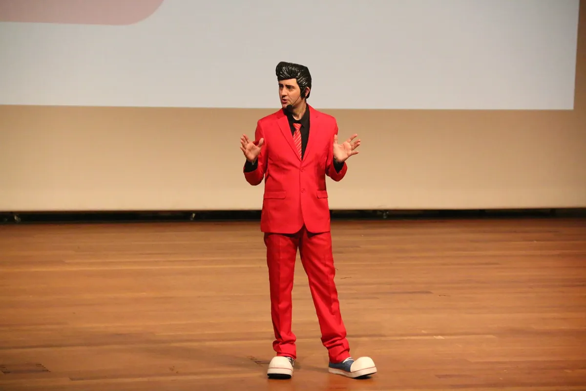 Apresentador vestido com terno vermelho e sapatos grandes fala ao microfone em palco de madeira diante de tela branca.