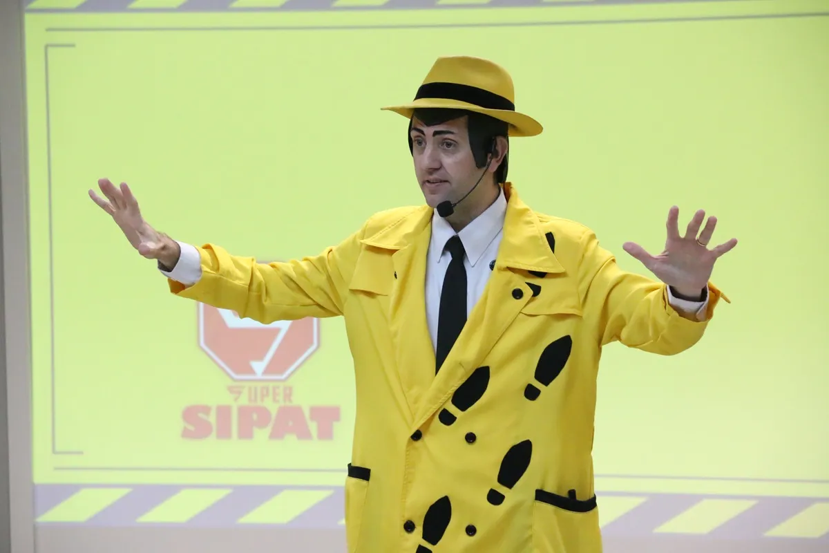 Facilitador de sobretudo amarelo pontilhado de pegadas gesticula em palestra de segurança diante de tela amarela com logo Super SIPAT.