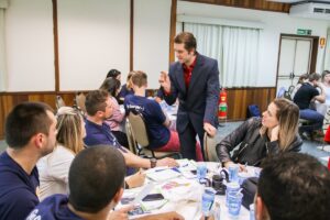 Local de trabalho com pessoas distribuídas em mesa enquanto palestrante fala em meio a essas pessoas durante discussão sobre Logo CIPA