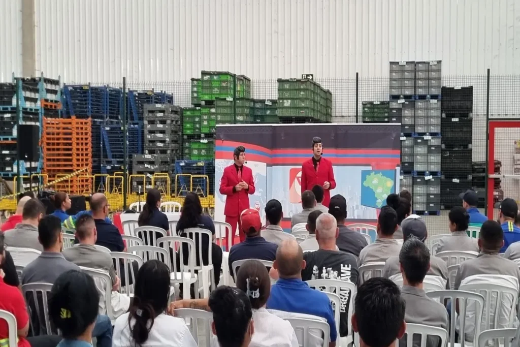 Dois palestrantes ou oradores vestindo ternos vermelhos em um palco simples, falando para uma plateia de funcionários em um ambiente industrial, ilustrando um treinamento de oratória ou curso de comunicação empresarial.