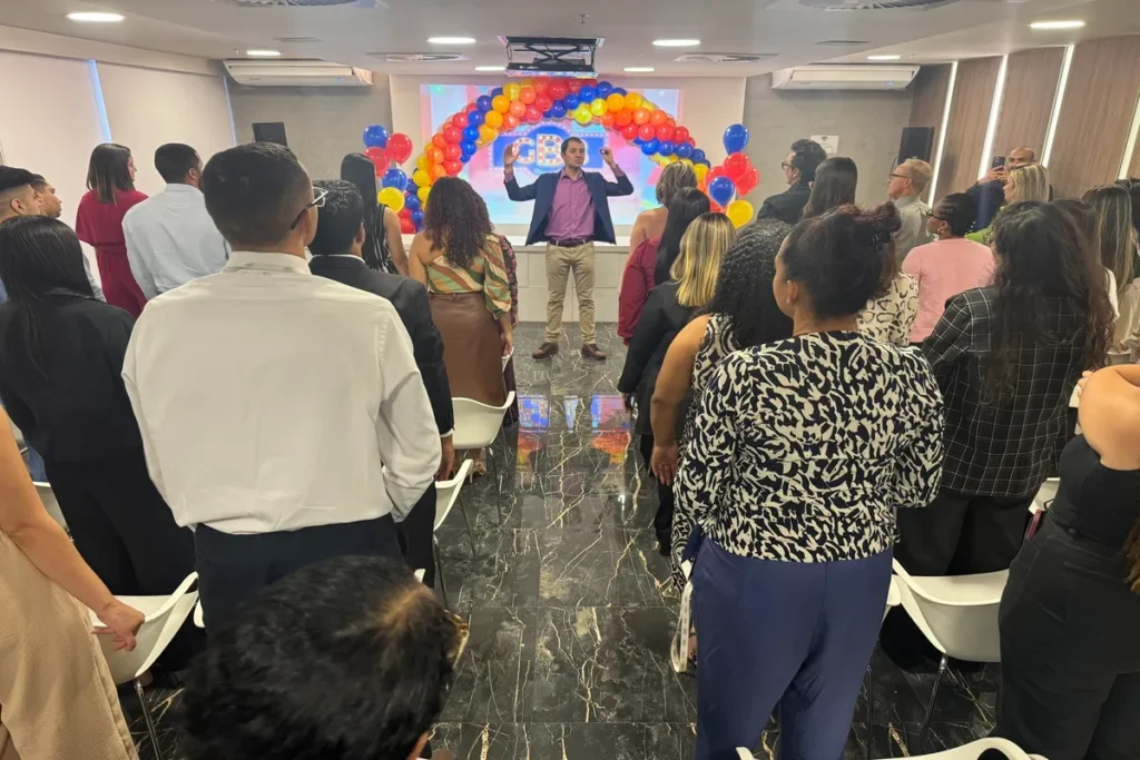 Um palestrante em pé no centro de uma sala de treinamento, gesticulando para um grupo de profissionais em um evento decorado com um arco de balões coloridos, simbolizando a importância da NR 10 e segurança no trabalho.