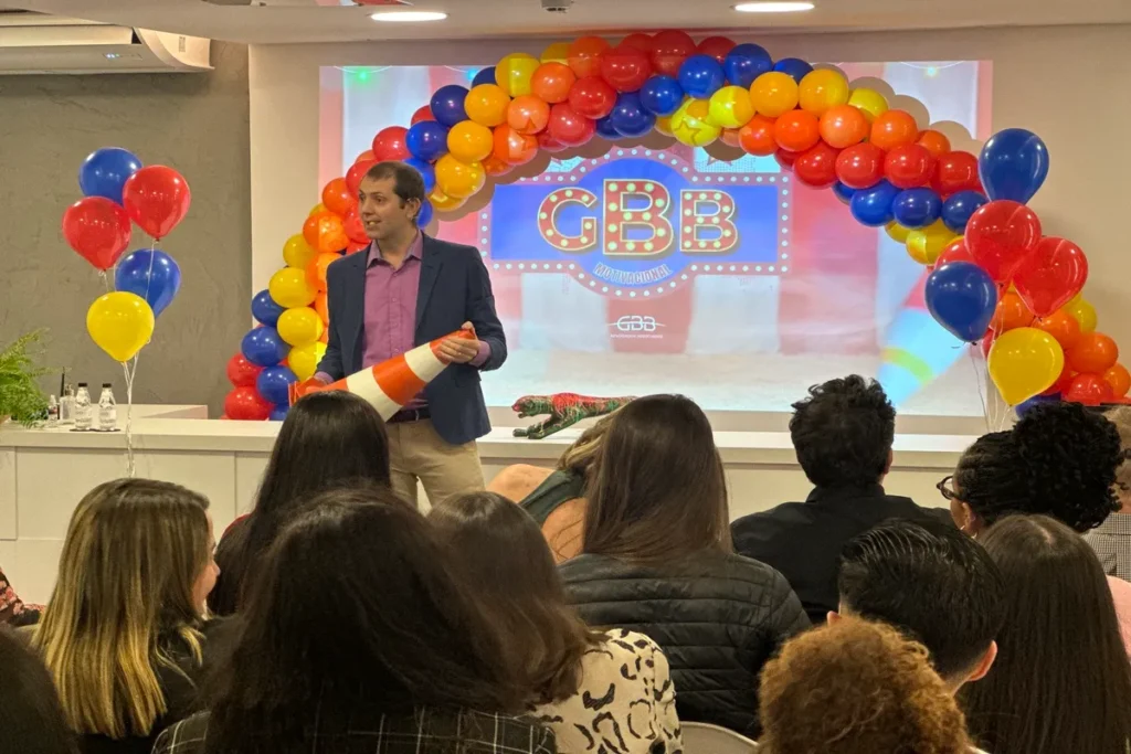 Um palestrante segurando um cone de trânsito em um palco de evento corporativo, com um grande arco de balões coloridos (vermelho, amarelo, azul) no fundo, dirigindo-se a uma plateia, ilustrando o treinamento de NR 11 e segurança na logística.