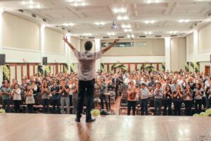Palestrante sobre palco de local próprio para eventos com braços erguidos e plateia aplaudindo diante do mesmo que fala sobre NR 15