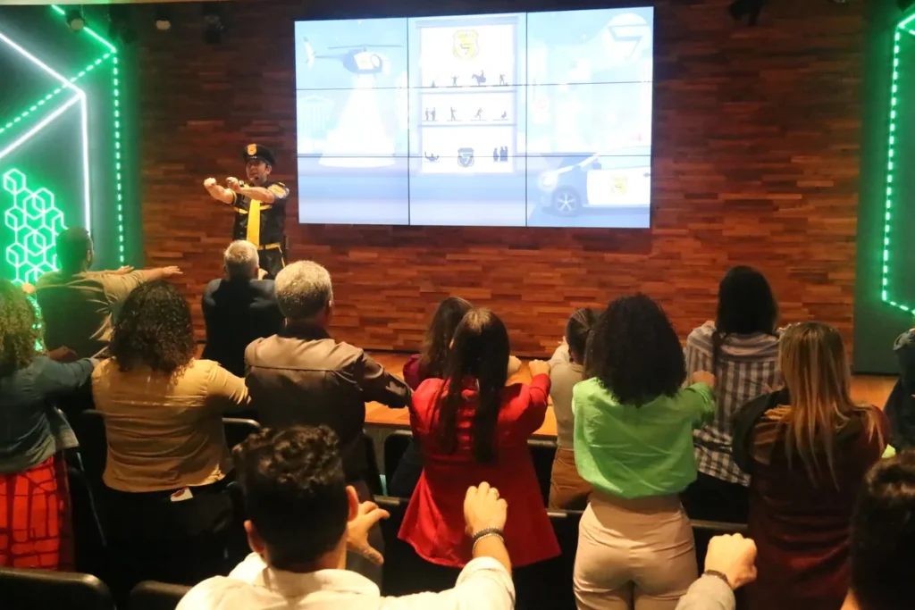 Um instrutor vestido como agente de segurança (em uniforme preto e amarelo) no palco de um auditório moderno, conduzindo uma dinâmica de grupo. A plateia (vista de costas) levanta os braços, engajada na atividade física ou exercício de conscientização sobre a NR 15 e segurança do trabalho.