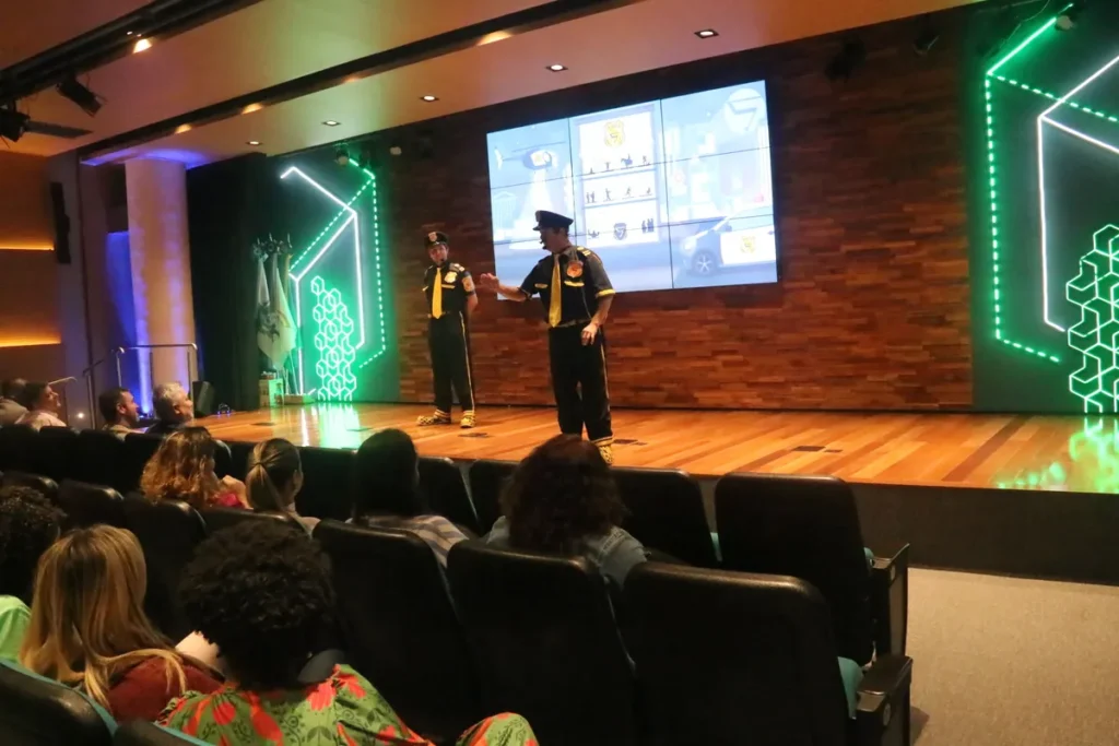 Dois palestrantes fantasiados de agentes de segurança (um com boné e outro com chapéu) em um palco de auditório, gesticulando em frente a uma tela de LED com gráficos de segurança. A cena, em um evento corporativo, ilustra o treinamento sobre a NR 15 e a prevenção de riscos insalubres.