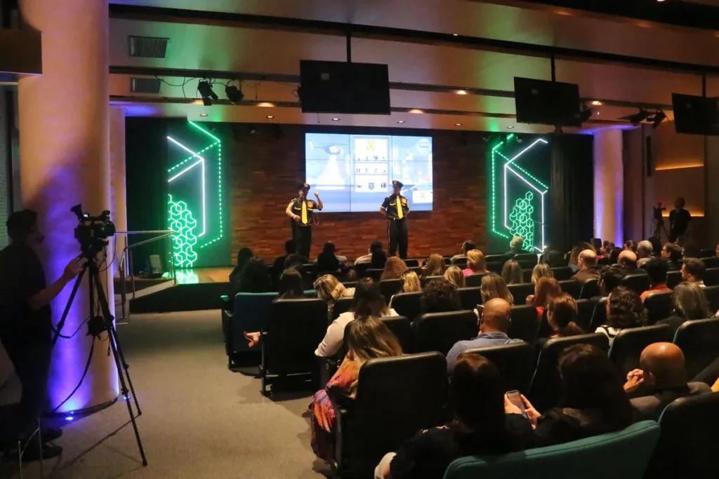 Dois palestrantes em uniformes de segurança no palco de um auditório com telões de LED, apresentando conteúdo sobre a NR 31 para uma grande plateia sentada, enquanto o evento é gravado por um cinegrafista.