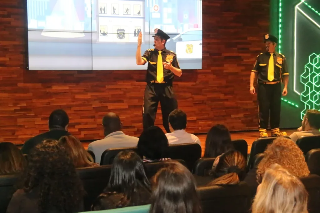 Dois palestrantes ou atores, vestidos com uniformes temáticos de segurança, se apresentam em um palco com um telão ao fundo para uma plateia, ilustrando um treinamento sobre a NR 31 de segurança no trabalho.