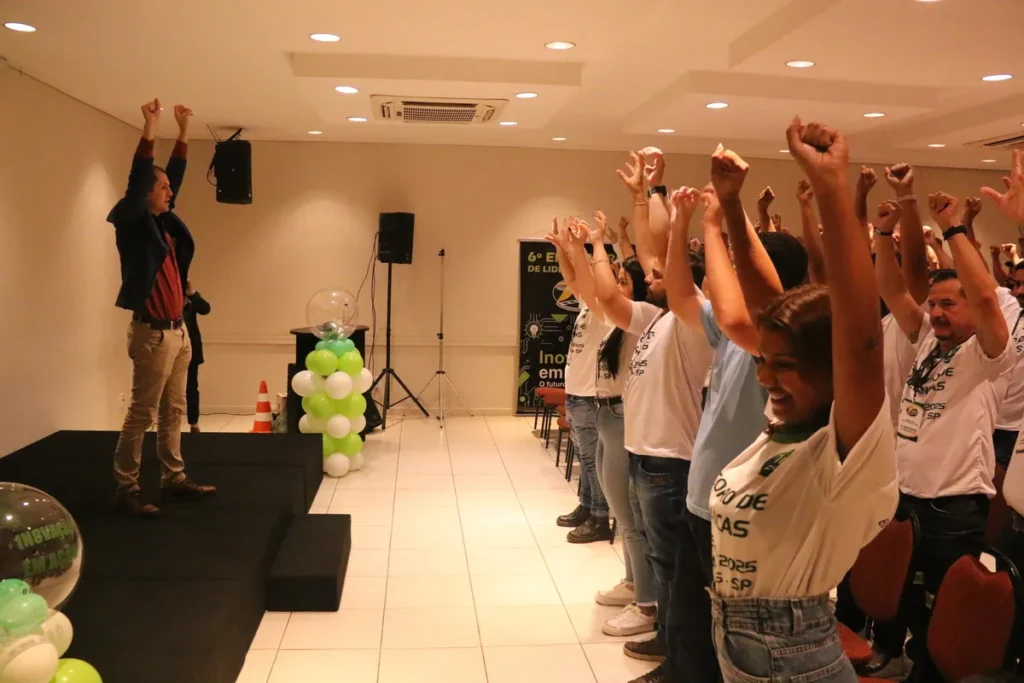Plateia em treinamento ou evento corporativo, com todos os participantes e um orador no palco erguendo os braços em um gesto de celebração ou motivação, representando o engajamento na CIPA (NR 5).