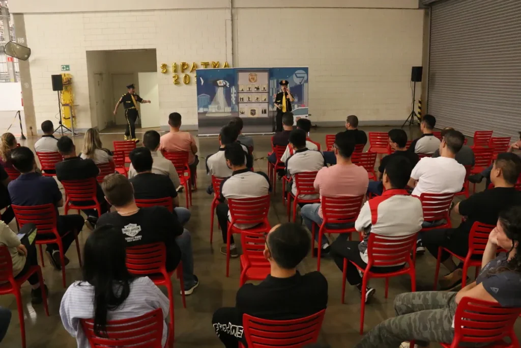Palestrante vestido como agente de segurança (camisa e boné amarelos) em uma SIPAT, apresentando em um palco a um público de colaboradores sentados em cadeiras vermelhas. O evento, com balões 'SIPATMA 2021', ilustra o treinamento sobre a NR 7 (PCMSO) e saúde ocupacional em ambiente industrial.