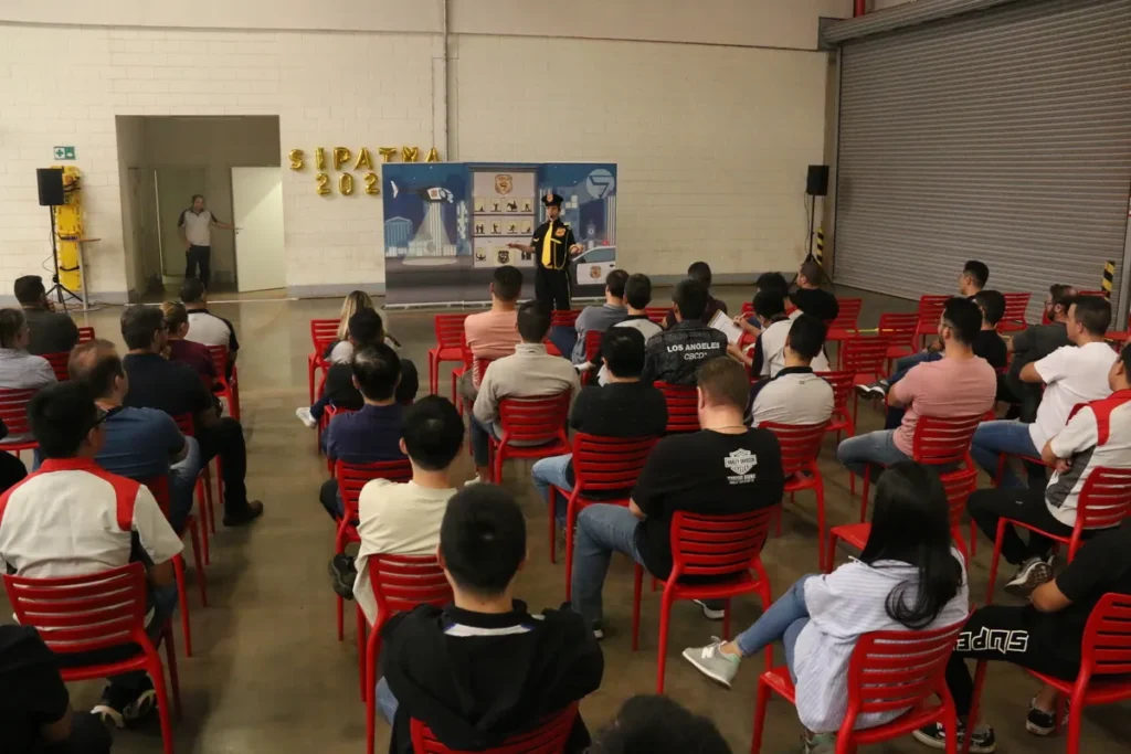 Uma plateia de colaboradores sentados em um auditório interno assiste a uma apresentação com slides no telão, com instrutores em pé ao lado. A cena, que mostra um treinamento interno com participantes vestindo uniformes escuros, representa a conscientização sobre a NR 7 e o PCMSO.
