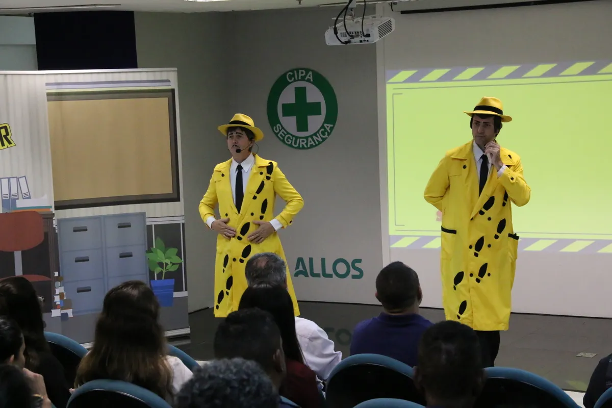 Dois homens vestidos como detetives ou inspetores (capas e chapéus amarelos com estampa de pegadas) em um palco de treinamento corporativo, com o logo da CIPA - Segurança ao fundo, representando uma palestra sobre a NR 9 e Prevenção de Riscos Ambientais (PPRA).