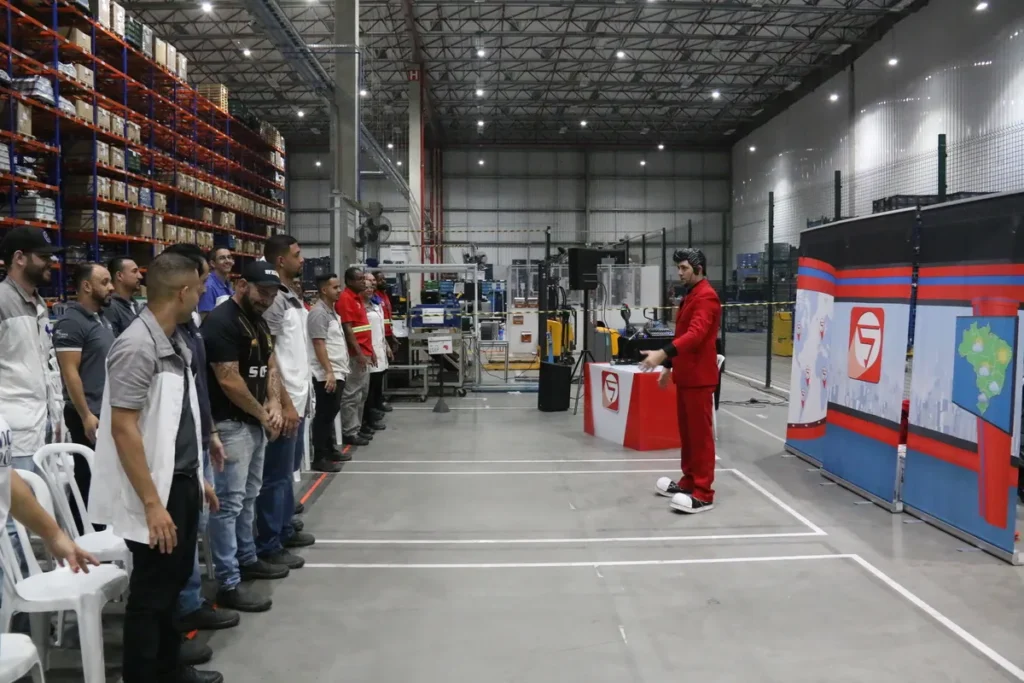Palestra ou treinamento em um galpão industrial, onde um homem de terno vermelho gesticula para uma fila de funcionários em uniformes, ilustrando a conscientização sobre o sedentarismo e a importância da saúde no ambiente de trabalho.